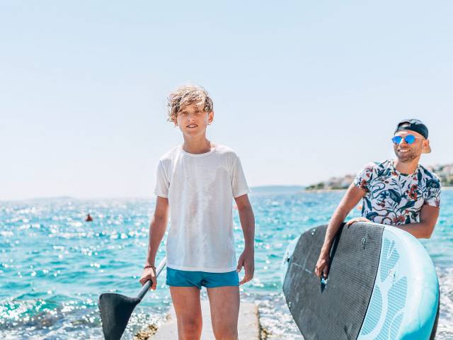 Junger Mann mit Cap und Surfbrett im Arm und Teenie mit Angelkelle in der Hand gehen vom Meer zum Strand