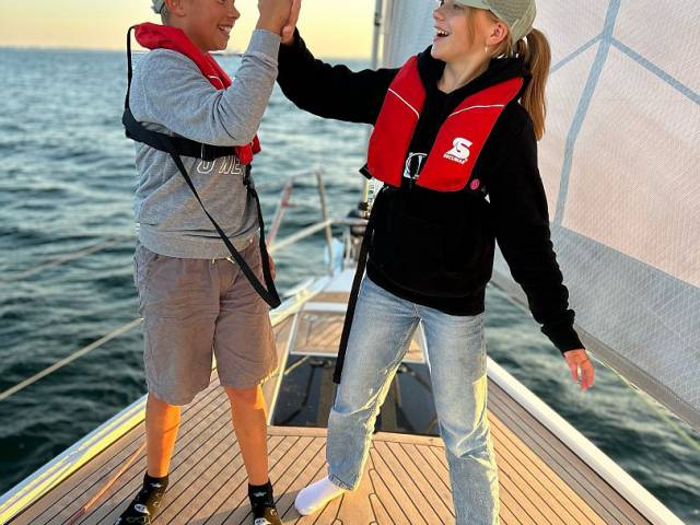 Mädchen und Junge mit Schwimmwesten & Caps klatschen sich auf dem Segelboot ab