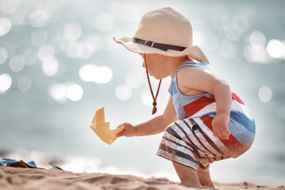 Kleinkind mit Hut spielt mit Schiffchen im Sand