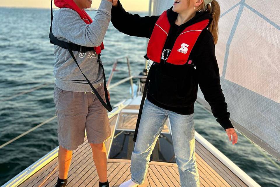 Mädchen und Junge mit Schwimmwesten & Caps klatschen sich auf dem Segelboot ab